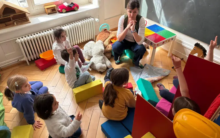 Vaspitačica sedi sa decom okupljenom u krug u igraonici, nekoliko dece diže ruku