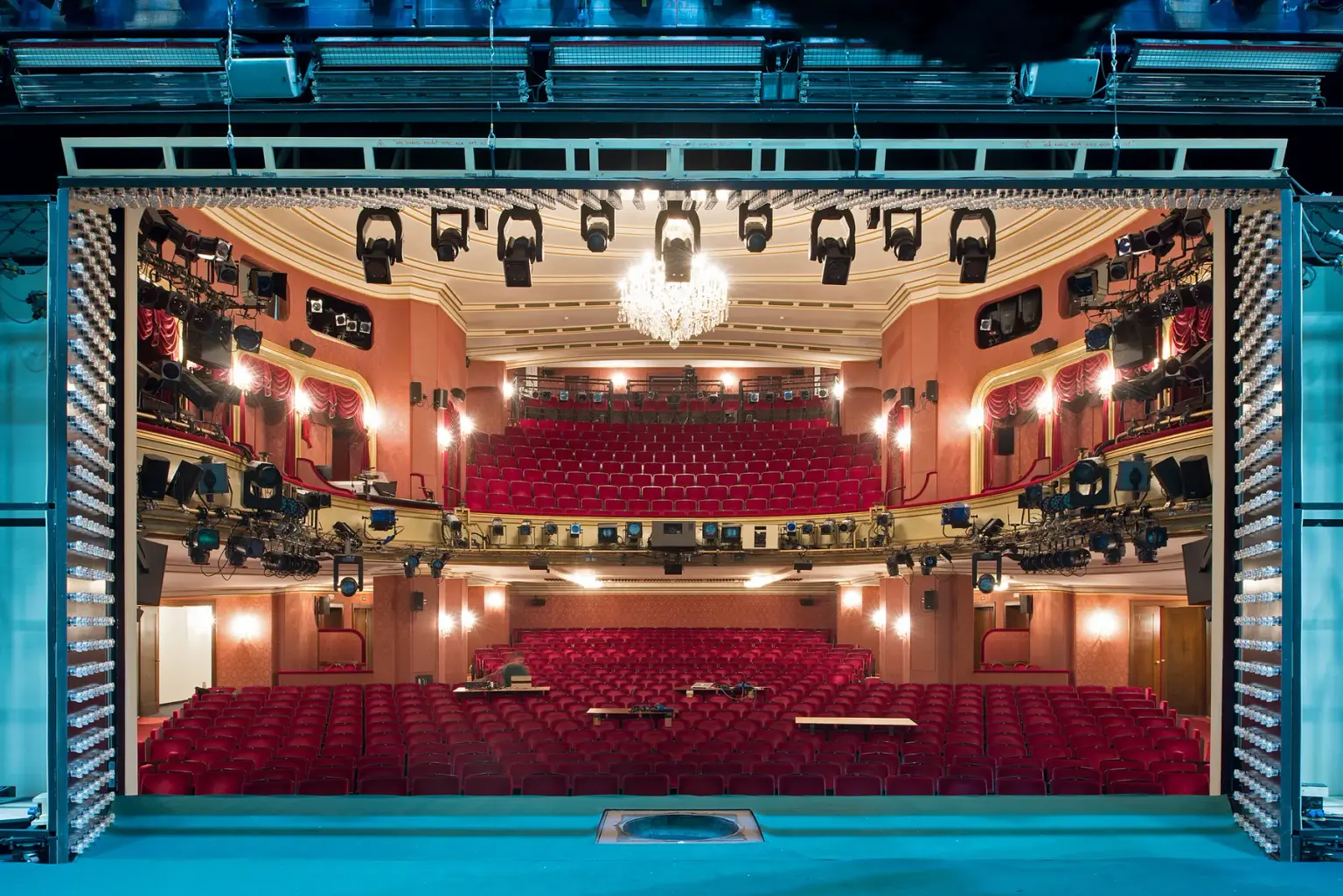 View from the stage through the proscenium into an empty theatre auditorium with red seats and a chandelier