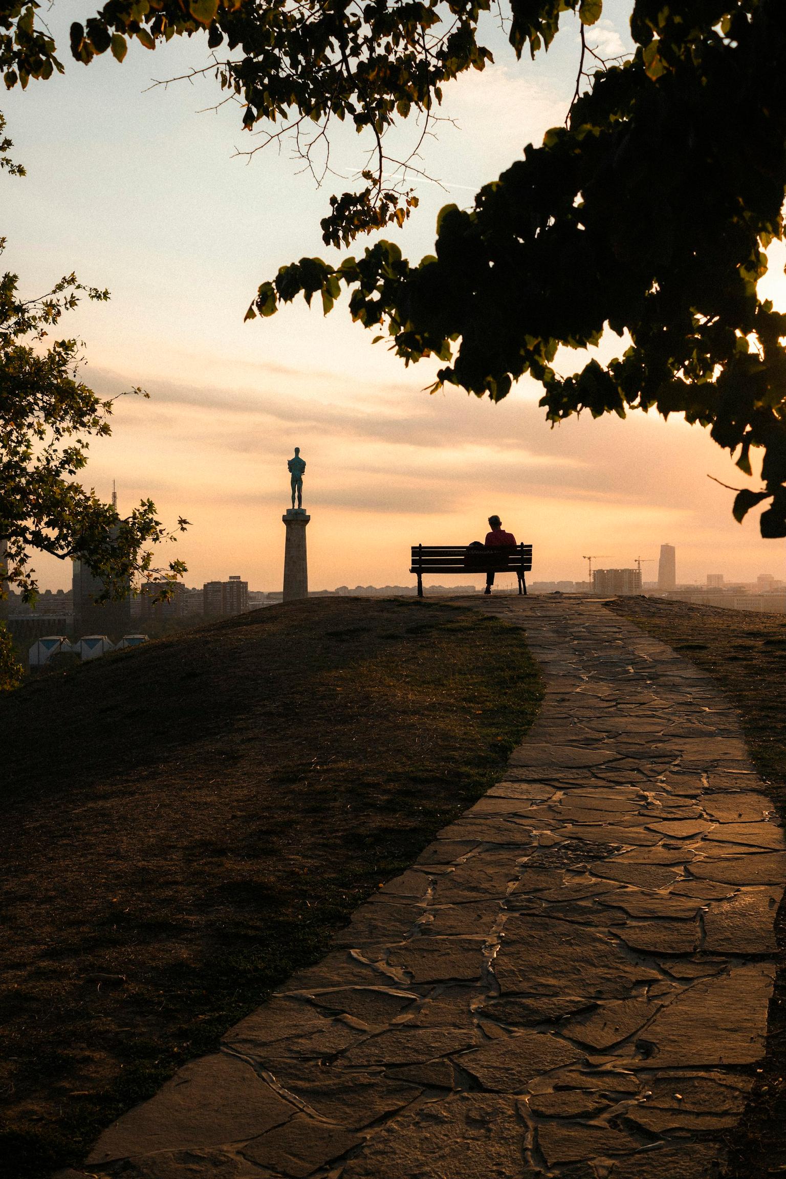 Silueta osobe na klupi na Kalemegdanu pored spomenika Pobedniku, sa pogledom na grad u sumrak